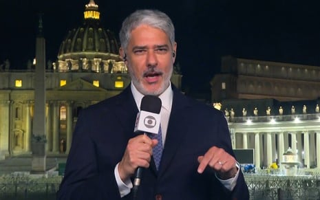 De terno e gravata, William Bonner segura microfone com a canopla da TV Globo na Praça São Pedro, no Vaticano