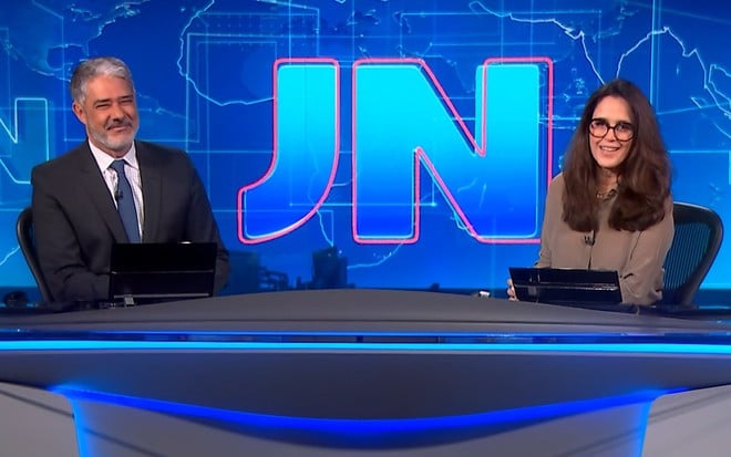 William Bonner ri na bancada do Jornal Nacional; Ana Luiza Guimarães está ao seu lado