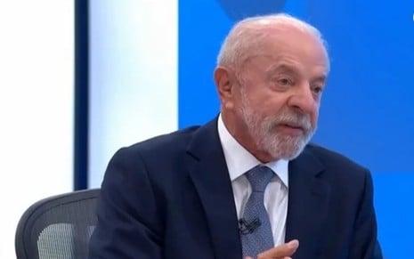 O presidente Luiz Inácio Lula da Silva surge em plano médio, de cabelo e barba grisalhos, vestindo um fato azul-escuro sobre uma camisa branca e gravata de padrão azul