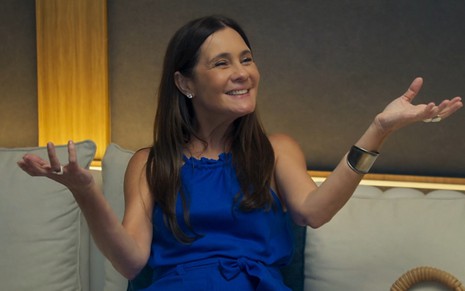 Sentada em um sofá, Adriana Esteves aparece sorridente em cena de Mania de Você