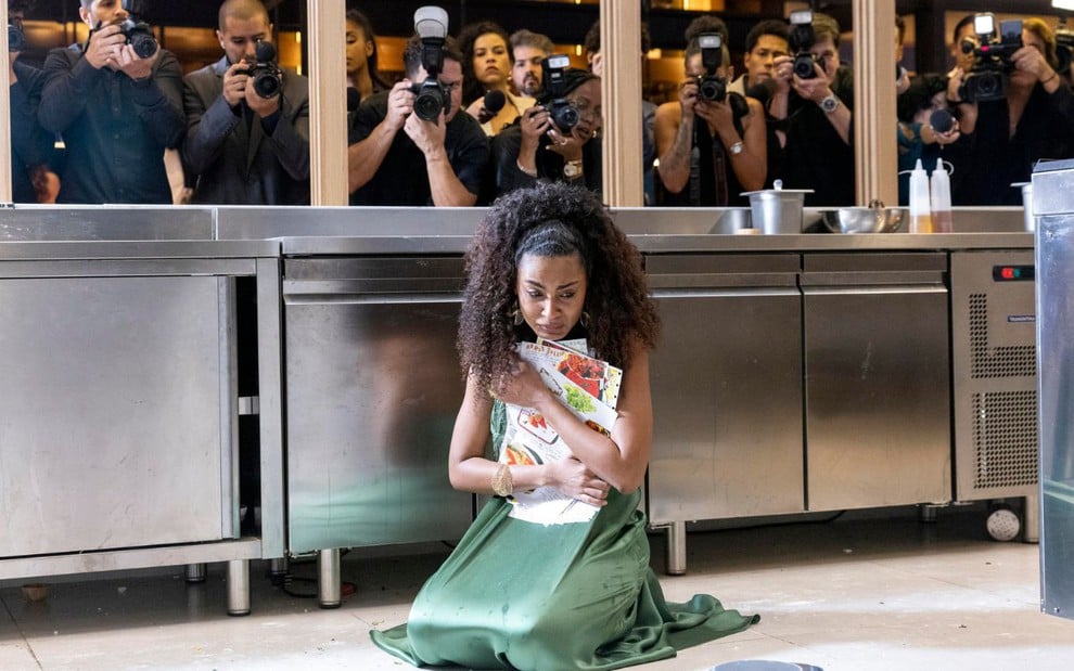 Uma mulher ajoelhada no chão, com expressão de tristeza, segura papéis em um cenário de cozinha industrial. Atrás, fotógrafos tiram fotos dela.