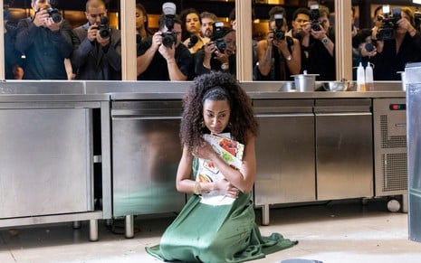 Uma mulher ajoelhada no chão, com expressão de tristeza, segura papéis em um cenário de cozinha industrial. Atrás, fotógrafos tiram fotos dela.