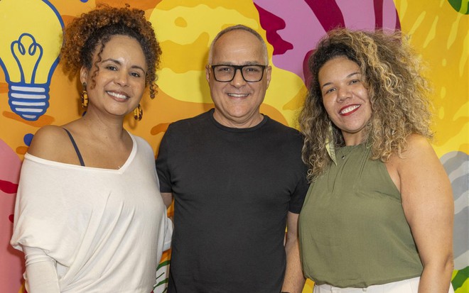 Renata Andrade, Amauri Soares e Thais Pontes posam sorridentes para foto em um backdrop colorido