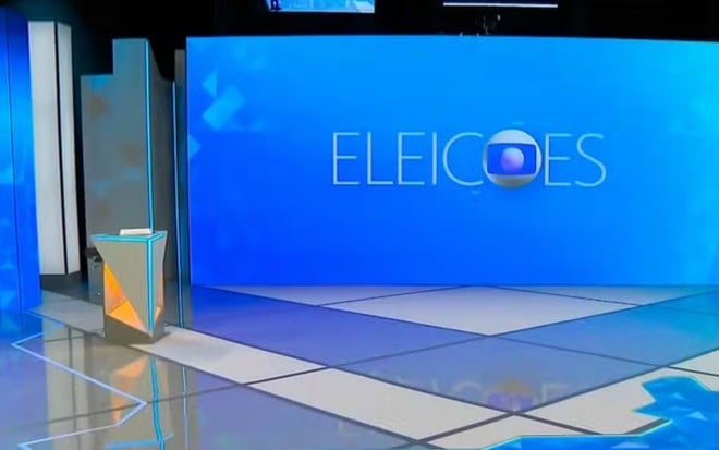 Cenário de estúdio de televisão preparado para cobertura eleitoral, com um grande painel azul ao fundo onde se lê a palavra "ELEIÇÕES" acompanhada pelo logótipo da TV Globo, e um púlpito iluminado em tons de cinzento e laranja no lado esquerdo.