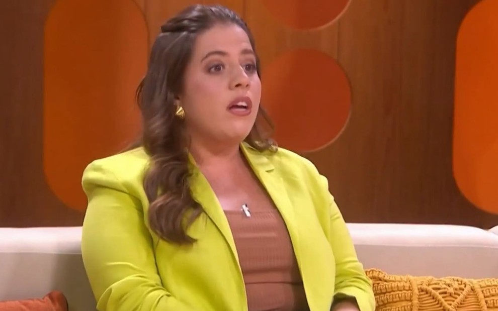 Tati Machado usa um top bege e um blazer amarelo; ela está boquiaberta no Saia Justa