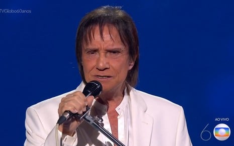 O cantor Roberto Carlos se apresenta de paletó e camisa branca diante de um fundo azul
