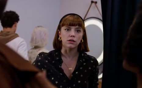 Mulher de expressão surpresa encara alguém; usa blusa preta com estrelas, tiara, brincos dourados e colar com nome “Bela”