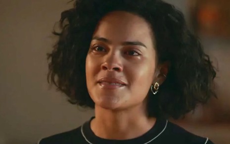 Mulher de cabelo cacheado curto aparece emocionada, com lágrimas nos olhos. Ela usa brincos dourados e blusa escura com detalhe claro na gola.        Perguntar ao ChatGPT