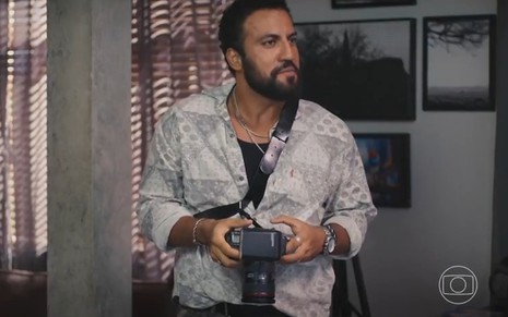 Ricardo Teodoro segura uma câmera fotográfica e tem expressão séria em cena de Vale Tudo