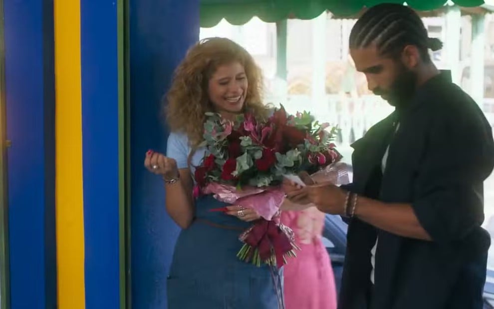 Isadora Cruz segura buquê de flores, sorridente, enquanto Vitor Sampaio parece contrariado