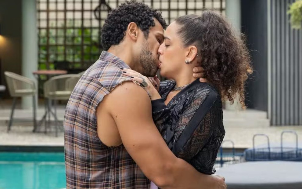 Um casal se beijando apaixonadamente em frente a uma piscina, com um ambiente moderno ao fundo. O homem usa uma camisa xadrez sem mangas, e a mulher, uma blusa preta com transparências.