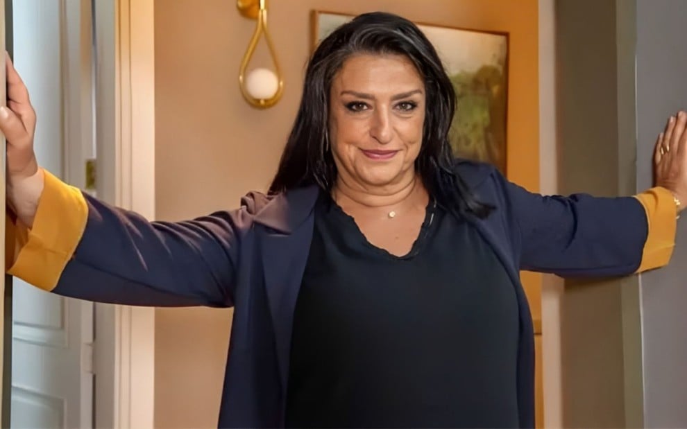Grace Gianoukas posando com as mãos apoiadas em um batente de portas no cenário de Família É Tudo (2024)