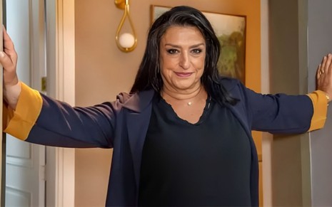 Grace Gianoukas posando com as mãos apoiadas em um batente de portas no cenário de Família É Tudo (2024)