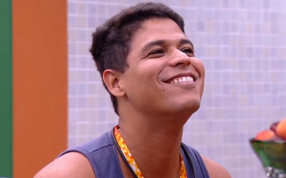 Guilherme Albuquerque sorridente no apê do líder no BBB 25; ele usa uma regata azul e está com colar laranja da liderança