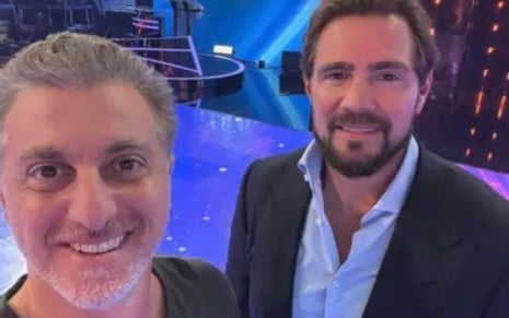 homem loiro e homem moreno barbado tiram selfie em estúdio de televisão