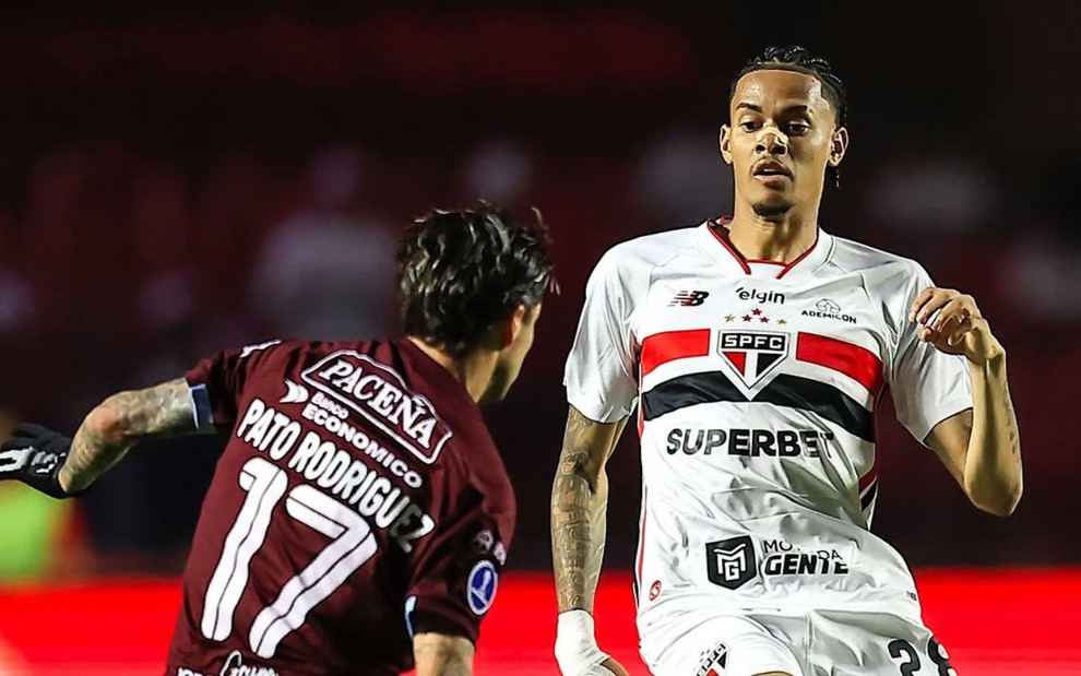 Newton do São Paulo, com curativo no nariz, conduz a bola em campo diante de Pato Rodríguez, vestindo camisa grená