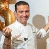 Buddy Valastro posa sorridente ao lado de bolo de várias camadas branco e vermelho