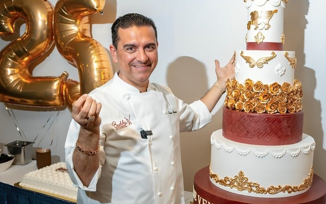 Buddy Valastro posa sorridente ao lado de bolo de várias camadas branco e vermelho