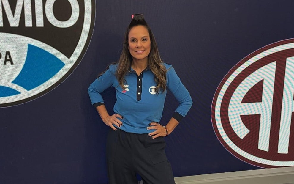 Vestindo uniforme da Globo, Fabiola Andrade posa com bobe no cabelo em frente a um telão