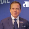João Doria posa com sorriso forçado em frente a um painel da Jovem Pan