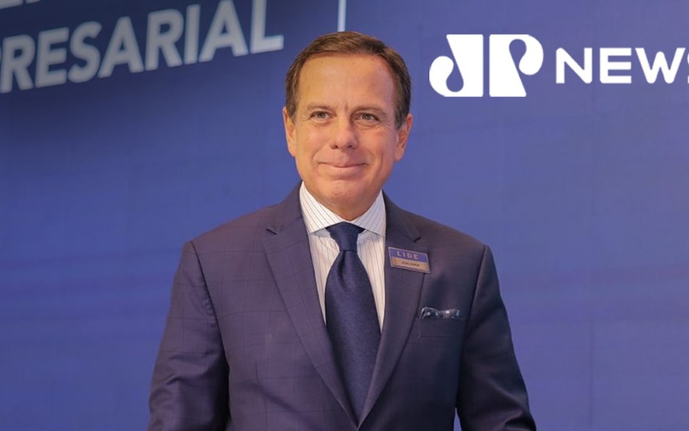 João Doria posa com sorriso forçado em frente a um painel da Jovem Pan