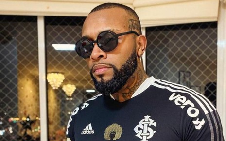 Nego Di posa na sala de uma casa com camisa de time de futebol e óculos escuros