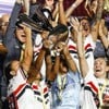 Mulheres com o uniforme do São Paulo levantam troféu em um palco montado no meio de um estádio de futebol