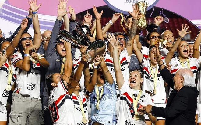 Mulheres com o uniforme do São Paulo levantam troféu em um palco montado no meio de um estádio de futebol