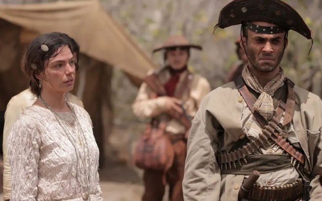 Homem com roupas de cangaceiro e mulher de vestido claro estão sérios em um cenário árido, com homens armados ao fundo.