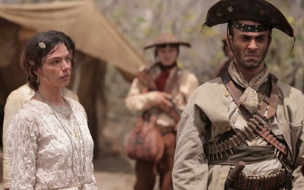 Homem com roupas de cangaceiro e mulher de vestido claro estão sérios em um cenário árido, com homens armados ao fundo.