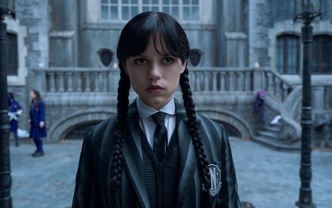 Jenna Ortega caracterizada como Wandinha, usando uniforme, tranças e feição séria