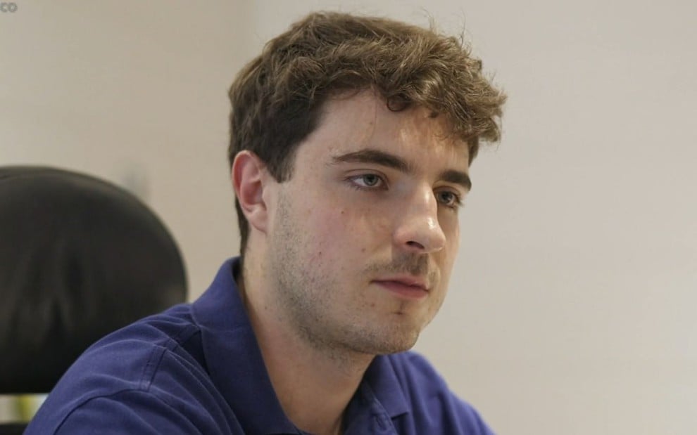 Homem jovem com expressão séria, cabelo castanho ondulado e camisa azul, sentado em ambiente interno com fundo neutro.