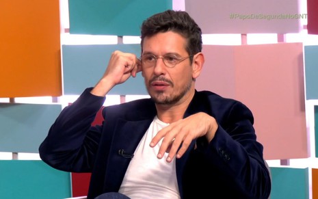João Vicente de Castro no Papo de Segunda, usando blazer, com uma mão segurando a orelha