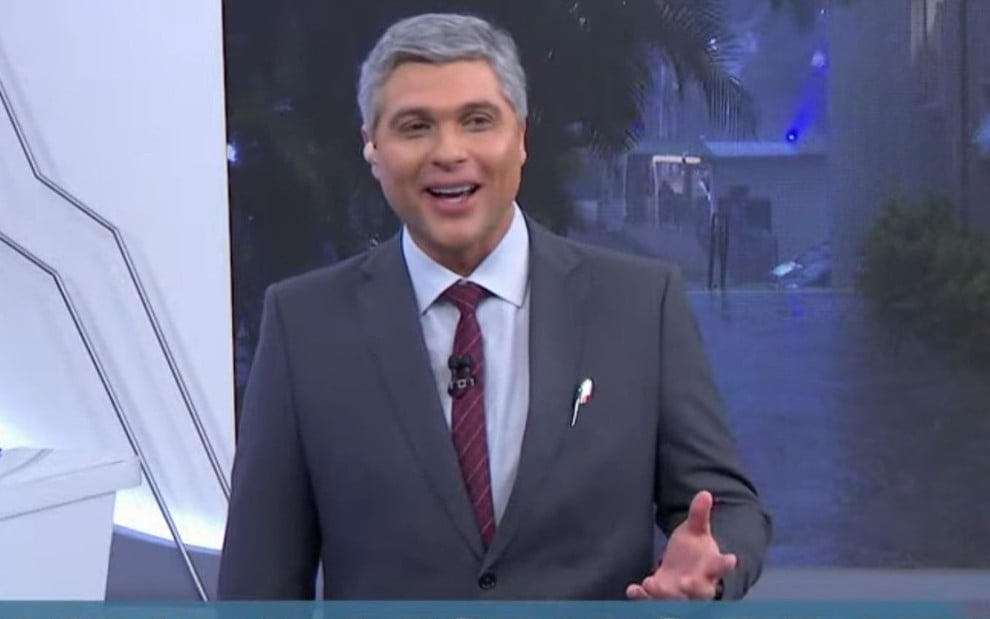 Apresentador de terno cinza e gravata vermelha, sorrindo em um estúdio de TV com fundo externo de cenário urbano à noite