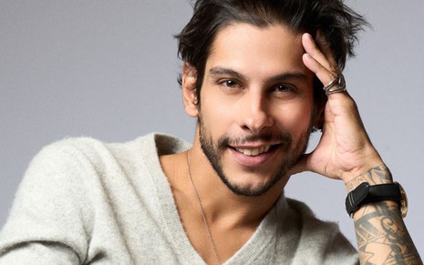 John Johny, irmão gêmeo de Shia Phoenix, posando com a mão na cabeça, sorridente, em um fundo cinza