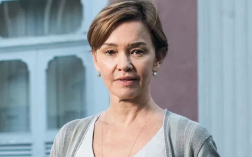 Julia Lemmertz olhando diretamente para a câmera, enquanto usa uma camiseta branca e um cardigan cinza