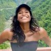 Mulher jovem sorri de braços abertos em ponte com vegetação densa ao fundo. Ela usa top preto e boné azul