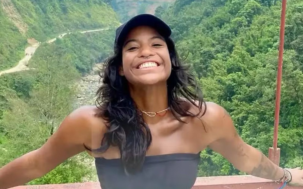 Mulher jovem sorri de braços abertos em ponte com vegetação densa ao fundo. Ela usa top preto e boné azul