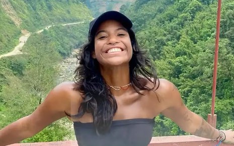 Mulher jovem sorri de braços abertos em ponte com vegetação densa ao fundo. Ela usa top preto e boné azul