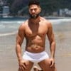 Montagem com fotos de Juninho e Nizam, do BBB 24. À esquerda, Juninho está ajoelhado de sunga branca na praia; à direita, Nizam posa sem camisa, tapando as partes íntimas com um pano