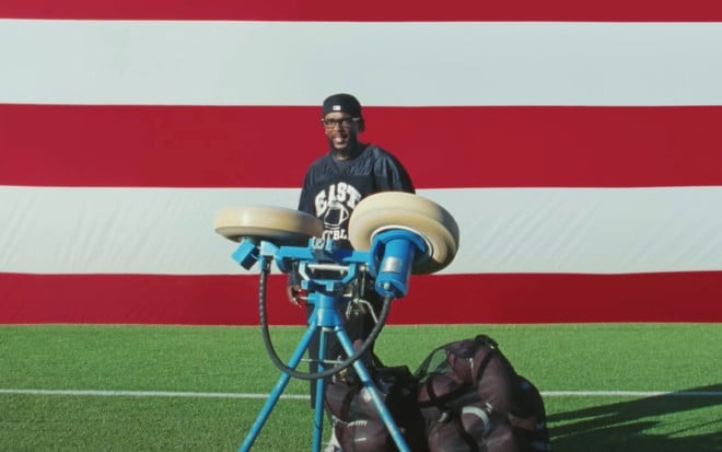 Kendrick Lamar em um estádio de futebol americano, com uma bandeira dos Estados Unidos ao fundo