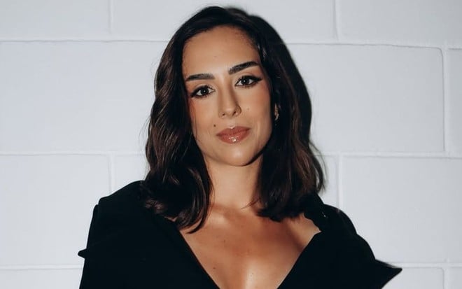 Cantora Lara posando com sorriso leve, vestido preto com colo à mostra, em frente a uma parede branca de tijolos
