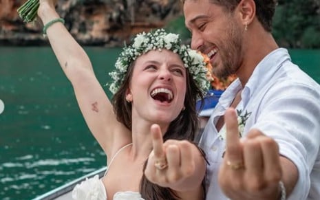 Larissa Manoela e André Luiz Frambach sorrindo em uma barco, vestidos de branco, e exibindo o anel de casamento