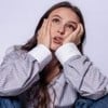 Larissa Manoela posando em fundo branco, com camisa social listrada, segurando o rosto com as mãos e uma expressão de aborrecimento