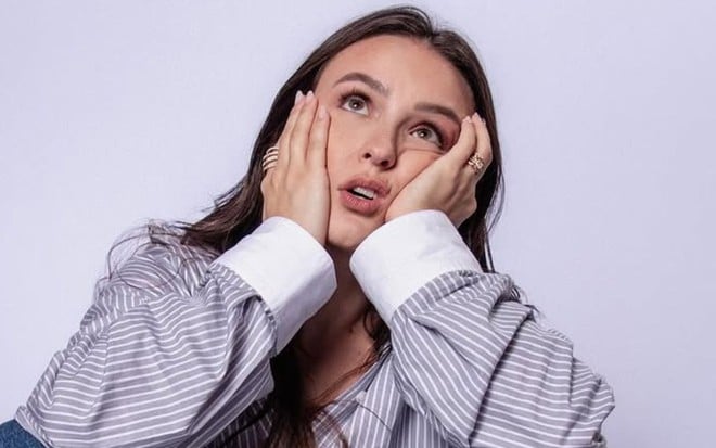 Larissa Manoela posando em fundo branco, com camisa social listrada, segurando o rosto com as mãos e uma expressão de aborrecimento