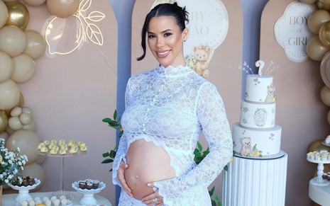 Larissa Tomásia usa uma roupa rendada branca que deixa à mostra a barriga de grávida; ao fundo estão itens de decoração em chá revelação