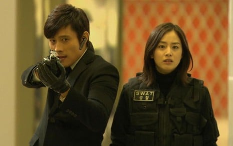 Lee Byung-hun aponta uma arma para frente, e Kim Tae-hee está com expressão preocupada em cena da série Iris