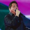 Leonardo em cima do palco, de camisa preta, cantando sorridente em frente a um fundo roxo