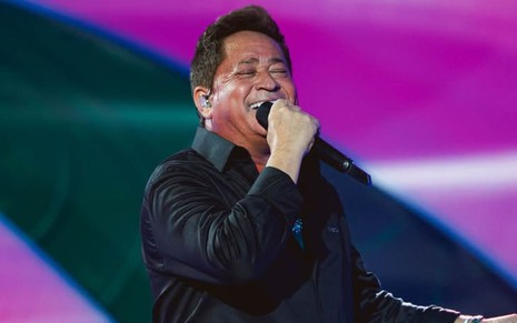 Leonardo em cima do palco, de camisa preta, cantando sorridente em frente a um fundo roxo