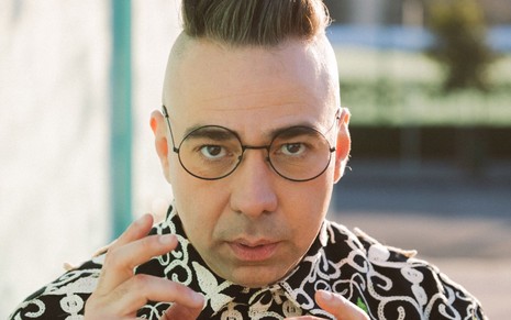 Ator de óculos redondos de armação preta e cabelo em estilo moicano, gesticula com as mãos ao olhar para a câmara